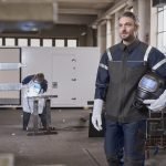 Un homme portant des vêtements de protection, notamment une veste de soudage et tenant un casque de soudage à obscurcissement automatique, se tient en toute confiance dans un espace de travail industriel, avec un autre travailleur en train de souder en arrière-plan.