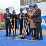 Un groupe de six personnes portant des casques bleus et des tenues formelles participent à une cérémonie d'inauguration des travaux. Ils tiennent chacun une pelle et se tiennent debout sur une bâche bleue. Une grande enseigne et plusieurs ouvriers en arrière-plan sont visibles.