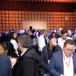 Un grand groupe de personnes, vêtues de tenues de travail, se rassemblent et conversent dans une salle de conférence bien éclairée dotée d'un mur à motifs géométriques. Les individus portent des badges nominatifs attachés à des cordons. Certaines personnes semblent concentrées sur leur téléphone ou leurs papiers.