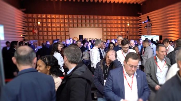 Un grand groupe de personnes, vêtues de tenues de travail, se rassemblent et conversent dans une salle de conférence bien éclairée dotée d'un mur à motifs géométriques. Les individus portent des badges nominatifs attachés à des cordons. Certaines personnes semblent concentrées sur leur téléphone ou leurs papiers.