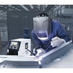 Un travailleur en tenue de protection effectue un contrôle de soudage en temps réel sur un composant métallique dans un environnement industriel, avec des étincelles volant autour.