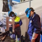 Deux travailleurs en tenue de protection effectuent un contrôle de soudage sur un composant métallique en milieu industriel, l'un observant et l'autre actionnant l'équipement.