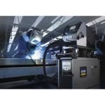 Un travailleur portant un casque de protection utilise une machine à souder sur une structure métallique dans un environnement industriel avec des étincelles visibles, des équipements sophistiqués et un contrôle qualité en temps réel.