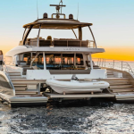 Un grand catamaran de luxe, ancré sur des eaux calmes au coucher du soleil, dispose de coins salons spacieux et d'un pont supérieur. A l'arrière, un petit canot blanc nommé "WILLIAMS" complète l'élégance. Dans la perspective des avancées de l'Industrie 4.0, une seule personne se tient sur le côté gauche du bateau.