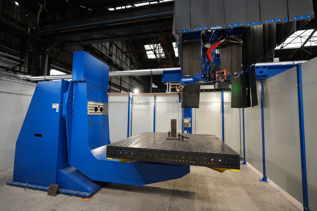 Une grande machine industrielle bleue à l’intérieur d’une installation spacieuse. La machine est dotée d'une large table plate avec une structure métallique au milieu et divers composants mécaniques au-dessus. La zone est bien éclairée, avec des murs, des tuyaux et des structures de plafond visibles en arrière-plan.