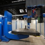 Une grande machine industrielle bleue à l’intérieur d’une installation spacieuse. La machine est dotée d'une large table plate avec une structure métallique au milieu et divers composants mécaniques au-dessus. La zone est bien éclairée, avec des murs, des tuyaux et des structures de plafond visibles en arrière-plan.
