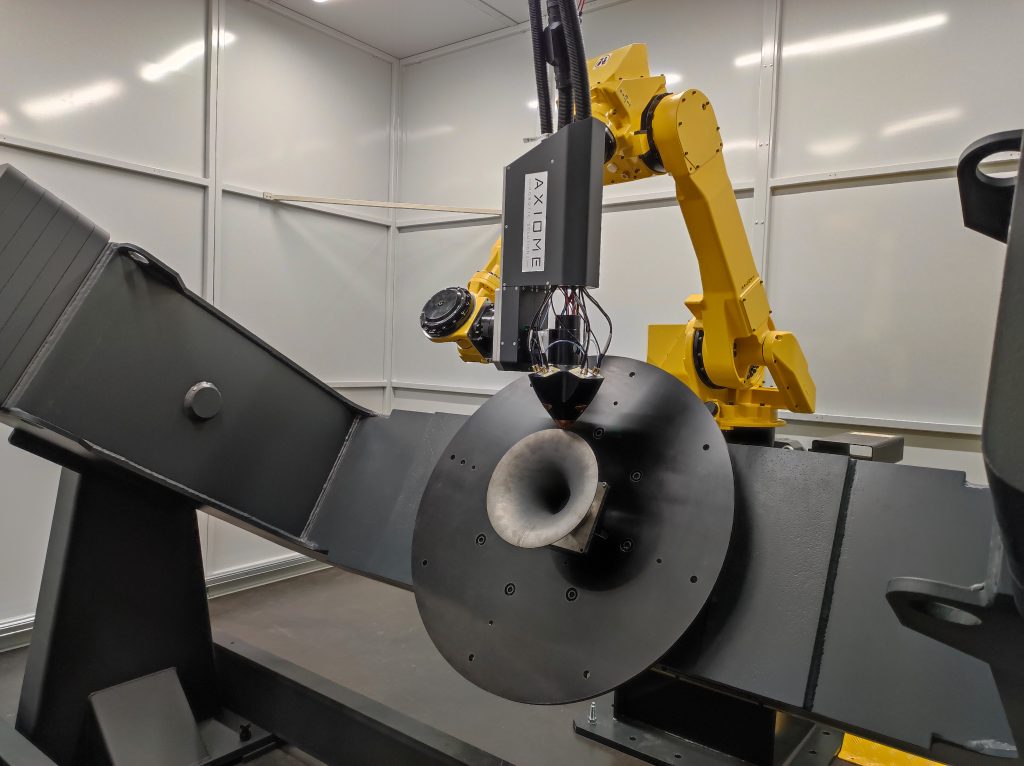 Un bras robotique jaune est vu en train de souder ou d'effectuer une tâche sur un composant métallique partiellement assemblé. L'installation se déroule dans une pièce industrielle bien éclairée avec des murs gris. L'équipement porte un étiquetage visible de la marque « AXIOME ».