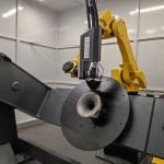 Un bras robotique jaune est vu en train de souder ou d'effectuer une tâche sur un composant métallique partiellement assemblé. L'installation se déroule dans une pièce industrielle bien éclairée avec des murs gris. L'équipement porte un étiquetage visible de la marque « AXIOME ».