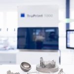 Une sélection d'objets métalliques imprimés en 3D sont exposés sur une table devant une imprimante 3D TruPrint 1000. Les objets comprennent une pièce sphérique, une structure en treillis et diverses pièces métalliques complexes. L'arrière-plan est légèrement flou, mettant en valeur les éléments imprimés.