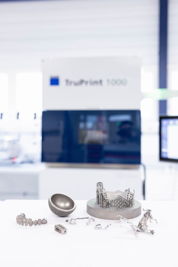 Une sélection d'objets métalliques imprimés en 3D sont exposés sur une table devant une imprimante 3D TruPrint 1000. Les objets comprennent une pièce sphérique, une structure en treillis et diverses pièces métalliques complexes. L'arrière-plan est légèrement flou, mettant en valeur les éléments imprimés.