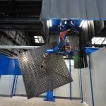 Une grande machine industrielle se trouve dans une usine. La machine est dotée d'un bras robotique tenant une tôle perforée. L'installation est bleue et grise, avec le nom de l'entreprise « Yaskawa » visible sur la machine. L’arrière-plan comprend de hauts plafonds et un éclairage industriel.