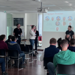 Un groupe de personnes est assis dans une salle de conférence de l'Institut de Soudure, face à un écran affichant une diapositive intitulée « Promesses des collègues ». Plusieurs individus se tiennent devant, parlant ou faisant une présentation. La salle dispose de grandes fenêtres laissant entrer la lumière naturelle, créant une atmosphère invitante pour cette visite immersive.