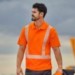 Un homme portant une chemise orange haute visibilité avec des bandes réfléchissantes se tient à l’extérieur, tenant une veste de couleur foncée. Le ciel nuageux forme l'arrière-plan, ainsi qu'une partie de la queue d'un avion, comme s'il venait de terminer une série de tests de sécurité sur des vêtements haute visibilité.