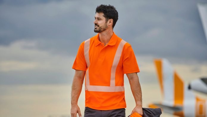 Un homme portant une chemise orange haute visibilité avec des bandes réfléchissantes se tient à l’extérieur, tenant une veste de couleur foncée. Le ciel nuageux forme l'arrière-plan, ainsi qu'une partie de la queue d'un avion, comme s'il venait de terminer une série de tests de sécurité sur des vêtements haute visibilité.