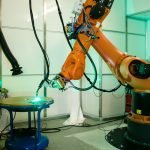Un bras robotique industriel opère dans un espace de travail bien éclairé, effectuant des tâches précises sur une table circulaire bleue. La scène comprend divers câbles et composants attachés au robot, reflétant l'adoption de techniques sophistiquées de configuration automatisée essentielles dans les environnements industriels modernes et la fabrication additive métallique.
