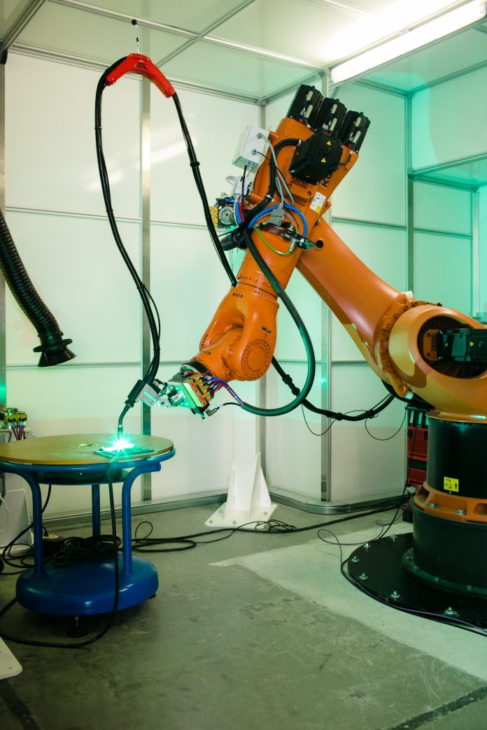 Un bras robotique industriel opère dans un espace de travail bien éclairé, effectuant des tâches précises sur une table circulaire bleue. La scène comprend divers câbles et composants attachés au robot, reflétant l'adoption de techniques sophistiquées de configuration automatisée essentielles dans les environnements industriels modernes et la fabrication additive métallique.