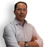 Un homme aux cheveux courts et portant des lunettes porte une chemise boutonnée de couleur claire. Il a les bras croisés et regarde directement la caméra, incarnant la précision d'un fabricant en France. Le fond est blanc uni.