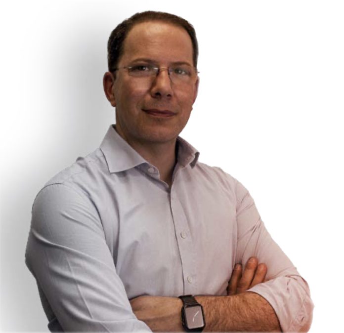 Un homme aux cheveux courts et portant des lunettes porte une chemise boutonnée de couleur claire. Il a les bras croisés et regarde directement la caméra, incarnant la précision d'un fabricant en France. Le fond est blanc uni.