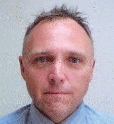 Un homme aux cheveux courts et hérissés et à l’expression sérieuse fait face à la caméra. Il porte une chemise bleu clair avec un signe facial EPI subtilement visible. Le fond est uni et blanc cassé.