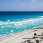 Une vue panoramique sur une plage aux eaux turquoise et au rivage de sable blanc. Les gens profitent de leurs vacances, se détendent sous des parasols en paille, se promènent le long du rivage et se baignent dans la mer. Le ciel est dégagé avec quelques nuages épars, tandis que les vagues se brisent doucement sur le rivage.