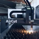 Une machine de découpe laser CNC baptisée « AirCut » fonctionne dans un atelier, émettant des étincelles brillantes lorsqu'elle découpe une feuille de métal sur une surface de support irrégulière. À proximité, un système d'extraction gère efficacement les débris, entouré d'équipements industriels et d'une unité de stockage.