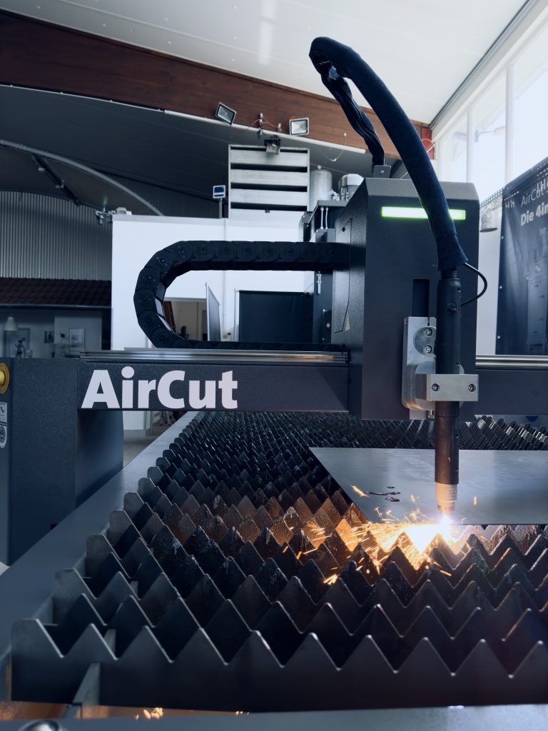Une machine de découpe laser CNC baptisée « AirCut » fonctionne dans un atelier, émettant des étincelles brillantes lorsqu'elle découpe une feuille de métal sur une surface de support irrégulière. À proximité, un système d'extraction gère efficacement les débris, entouré d'équipements industriels et d'une unité de stockage.