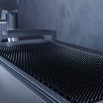 Machine industrielle avec une grande surface plane recouverte de pointes régulièrement espacées, probablement utilisées pour des solutions de découpe. L'équipement est placé contre un mur de béton gris uni, suggérant un décor d'usine ou d'atelier. La scène est brillamment éclairée, mettant en valeur les textures métalliques.