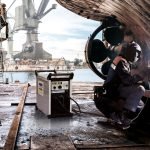 Un soudeur en tenue de protection effectue avec brio une soudure sur une structure métallique dans un chantier naval animé. À proximité, une machine à souder repose sur le sol. À l'arrière-plan, on aperçoit une grande grue et des bâtiments industriels au bord de l'eau, encadrés par un ciel partiellement nuageux.