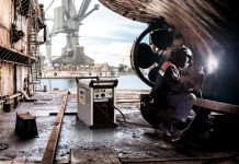 Un soudeur en tenue de protection effectue avec brio une soudure sur une structure métallique dans un chantier naval animé. À proximité, une machine à souder repose sur le sol. À l'arrière-plan, on aperçoit une grande grue et des bâtiments industriels au bord de l'eau, encadrés par un ciel partiellement nuageux.