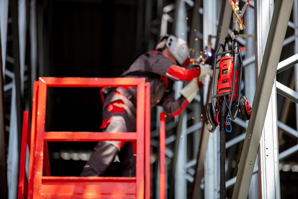 Une personne portant un équipement de protection exécute avec brio des soudures sur une structure métallique, debout sur un ascenseur rouge. Des étincelles jaillissent autour de la zone de soudage, avec une machine à souder rouge à proximité. Des poutres métalliques forment le décor, rappelant les chantiers navals animés.