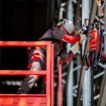 Une personne portant un équipement de protection exécute avec brio des soudures sur une structure métallique, debout sur un ascenseur rouge. Des étincelles jaillissent autour de la zone de soudage, avec une machine à souder rouge à proximité. Des poutres métalliques forment le décor, rappelant les chantiers navals animés.