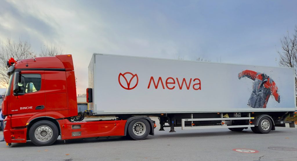 Un camion rouge avec une remorque blanche arbore le logo « Mewa ». Une image d'un t-shirt faisant la promotion des produits durables EPI apparaît sur la remorque. Le camion est garé sur une route bordée d'arbres sous un ciel nuageux, alliant harmonieusement la tranquillité de la nature à la qualité durable.