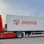 Un camion rouge avec une remorque blanche arbore le logo « Mewa ». Une image d'un t-shirt faisant la promotion des produits durables EPI apparaît sur la remorque. Le camion est garé sur une route bordée d'arbres sous un ciel nuageux, alliant harmonieusement la tranquillité de la nature à la qualité durable.