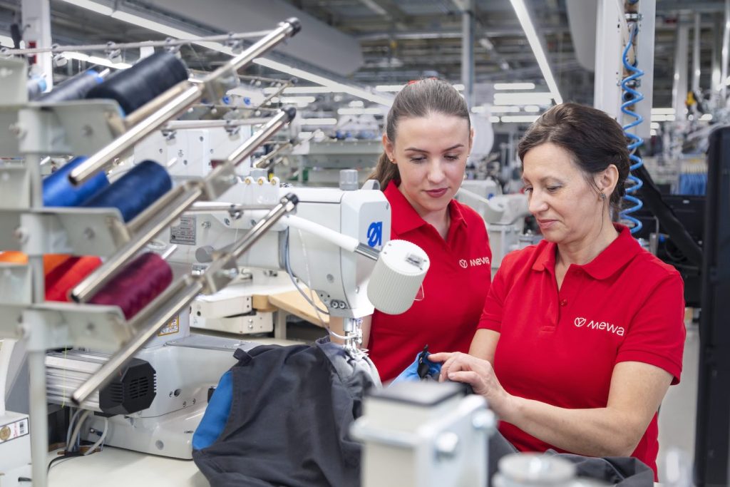 Deux femmes en chemises rouges travaillent ensemble dans une usine textile, entourées de machines à coudre et de bobines de fil colorées. Elles semblent concentrées alors qu'elles manipulent un morceau de tissu résistant, s'assurant que chaque détail est méticuleusement contrôlé pour la qualité.