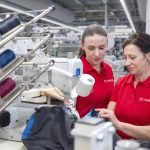 Deux femmes en chemises rouges travaillent ensemble dans une usine textile, entourées de machines à coudre et de bobines de fil colorées. Elles semblent concentrées alors qu'elles manipulent un morceau de tissu résistant, s'assurant que chaque détail est méticuleusement contrôlé pour la qualité.