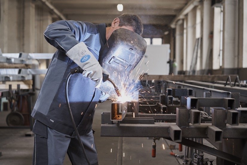 Une personne portant un équipement de protection soude du métal dans un atelier rempli de biens durables EPI. Des étincelles jaillissent lorsque la torche contrôlée entre en contact, illuminant le décor industriel avec ses sols et murs en béton, sur fond d'équipements et de poutres métalliques.