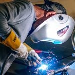 Une personne portant un casque de protection et des gants soude habilement une pièce de métal, des étincelles brillantes illuminant la scène industrielle. Positionné à gauche, le soudeur se concentre intensément sur son travail. Une petite lampe, suggérant la précision et le dévouement, est partiellement visible sur le côté droit.