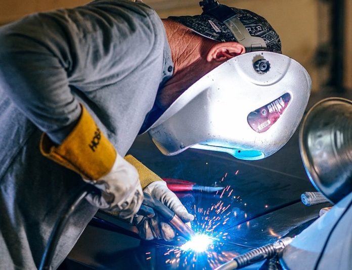 Une personne portant un casque de protection et des gants soude habilement une pièce de métal, des étincelles brillantes illuminant la scène industrielle. Positionné à gauche, le soudeur se concentre intensément sur son travail. Une petite lampe, suggérant la précision et le dévouement, est partiellement visible sur le côté droit.