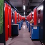 Un long couloir avec des rangées de cabines de soudage industrielles, chacune en lice pour le Championnat de France de Soudure, de chaque côté, séparées par des rideaux rouges. Les postes de soudage contiennent des machines bleues. Une bannière verte avec du texte et des graphiques blancs indistincts est visible au premier plan tandis que des plafonniers illuminent l'espace.