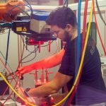 Un homme utilise des machines dans un environnement industriel, ajustant un équipement laser sous des lumières rouges et bleues avec divers câbles et fils visibles autour de l'espace de travail.