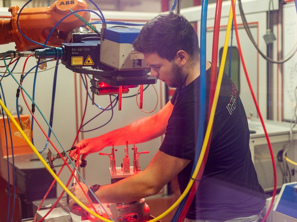 Un homme utilise des machines dans un environnement industriel, ajustant un équipement laser sous des lumières rouges et bleues avec divers câbles et fils visibles autour de l'espace de travail.