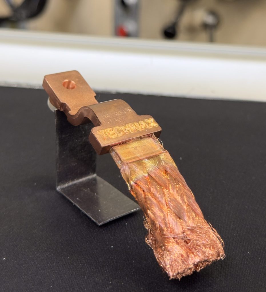 Un composant électrique en cuivre étiqueté « TECHMAX » est monté sur un support métallique, avec une extrémité tressée effilochée en bas, probablement traitée au laser. L'arrière-plan reste flou.