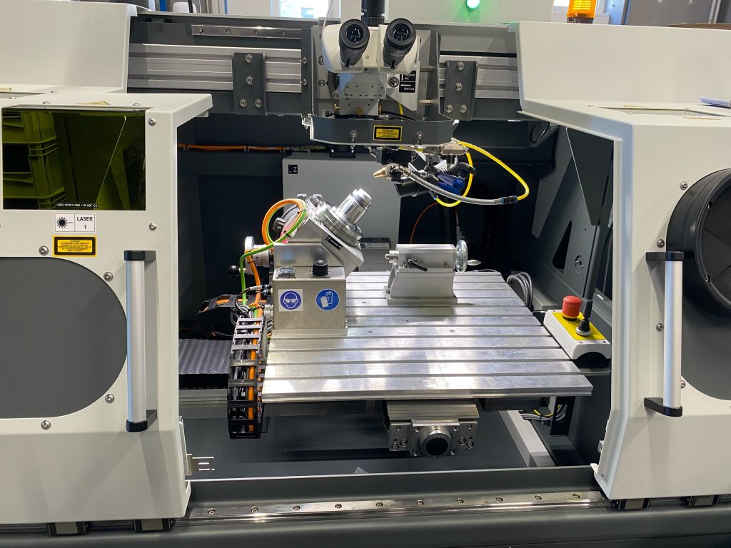 Une vue rapprochée de l'intérieur d'une machine CNC montrant une table de travail métallique, un axe rotatif, un câblage, des capteurs et des composants de contrôle, avec des étiquettes d'avertissement de sécurité et des boutons d'arrêt d'urgence, met en évidence l'intégration avancée de la technologie laser.