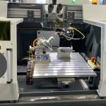 Une vue rapprochée de l'intérieur d'une machine CNC montrant une table de travail métallique, un axe rotatif, un câblage, des capteurs et des composants de contrôle, avec des étiquettes d'avertissement de sécurité et des boutons d'arrêt d'urgence, met en évidence l'intégration avancée de la technologie laser.