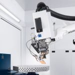 Un bras robotique industriel blanc étiqueté « KUKA » fonctionne à l'intérieur d'un espace de travail propre et blanc, utilisant un laser procédé alors qu'il plane au-dessus d'une plate-forme métallique avec des trous circulaires, entourée d'armoires et d'équipements.