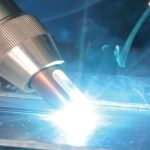 Gros plan d'un chalumeau de soudage émettant un arc lumineux brillant lors de l'assemblage de pièces métalliques, illustrant la précision de la technologie laser, avec des étincelles visibles et une légère brume de fumée en arrière-plan.