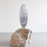 Une sculpture en métal de forme ovale, rappelant les techniques de Clara Imbert, avec de petites formes découpées, se dresse verticalement au sommet d'un gros rocher rugueux brun rougeâtre sur un sol gris sur un fond blanc uni.