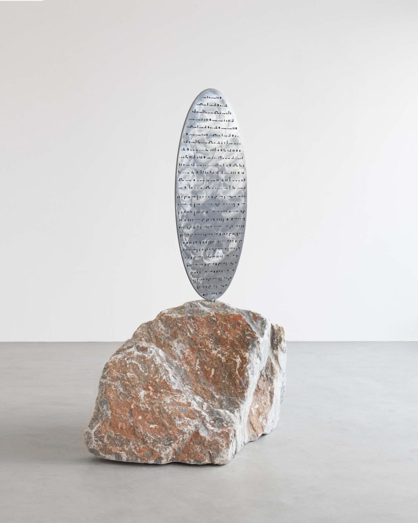 Une sculpture en métal de forme ovale, rappelant les techniques de Clara Imbert, avec de petites formes découpées, se dresse verticalement au sommet d'un gros rocher rugueux brun rougeâtre sur un sol gris sur un fond blanc uni.