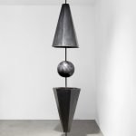 Une sculpture verticale de Clara Imbert présente des techniques intrigantes : une sphère noire nichée entre un cône inversé au-dessus et un cône vertical en dessous, le tout équilibré sur une tige fine au sommet d'une base circulaire dans une pièce blanche unie.