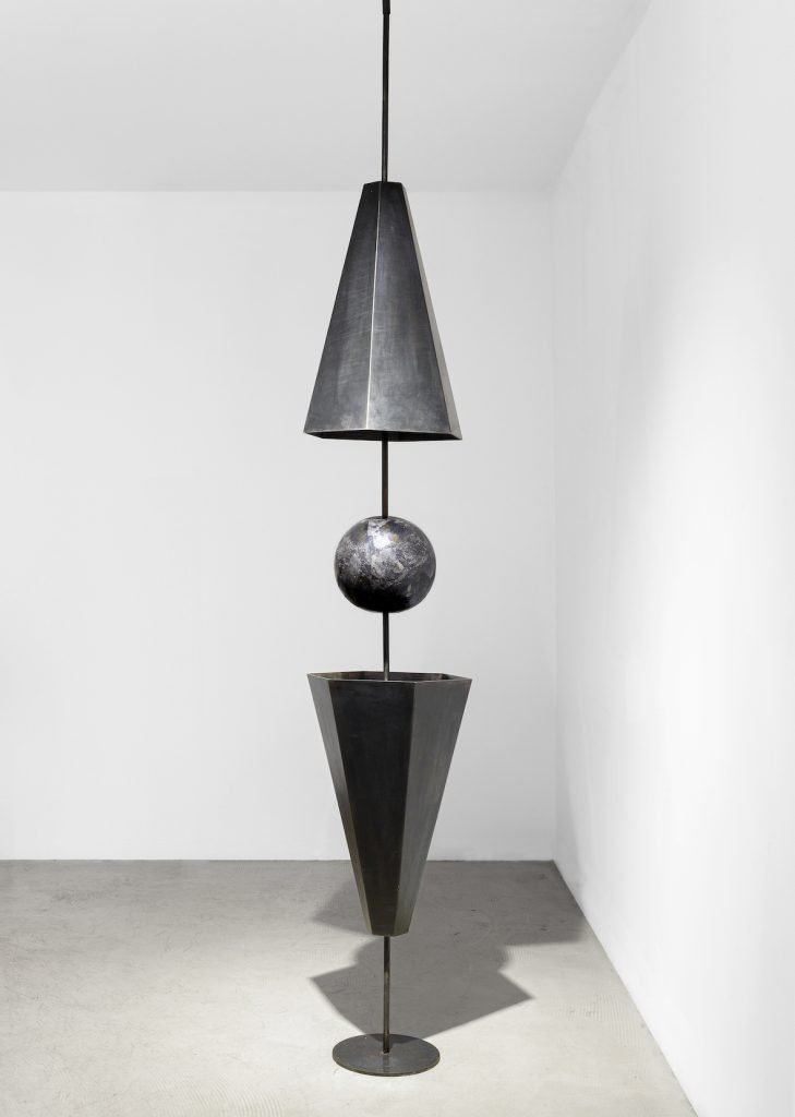Une sculpture verticale de Clara Imbert présente des techniques intrigantes : une sphère noire nichée entre un cône inversé au-dessus et un cône vertical en dessous, le tout équilibré sur une tige fine au sommet d'une base circulaire dans une pièce blanche unie.